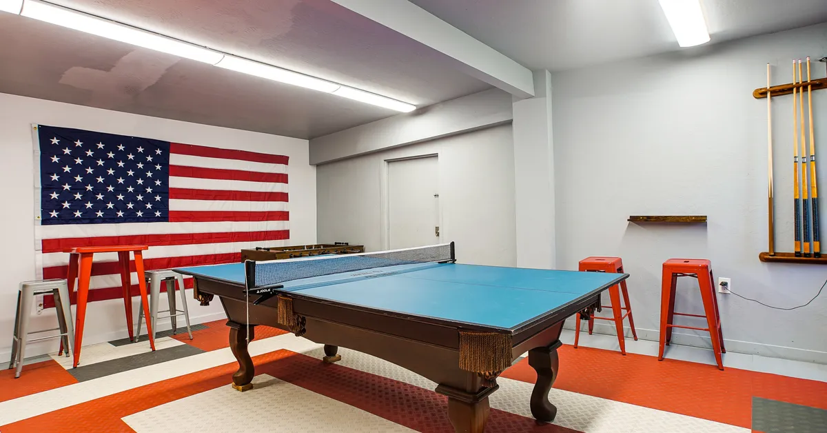 A well-equipped game room interior featuring a ping pong table, bar stools, and pool cues. An American flag decorates the wall, and the setup is inviting for both playing and spectating. The open space encourages camaraderie and group tournaments.