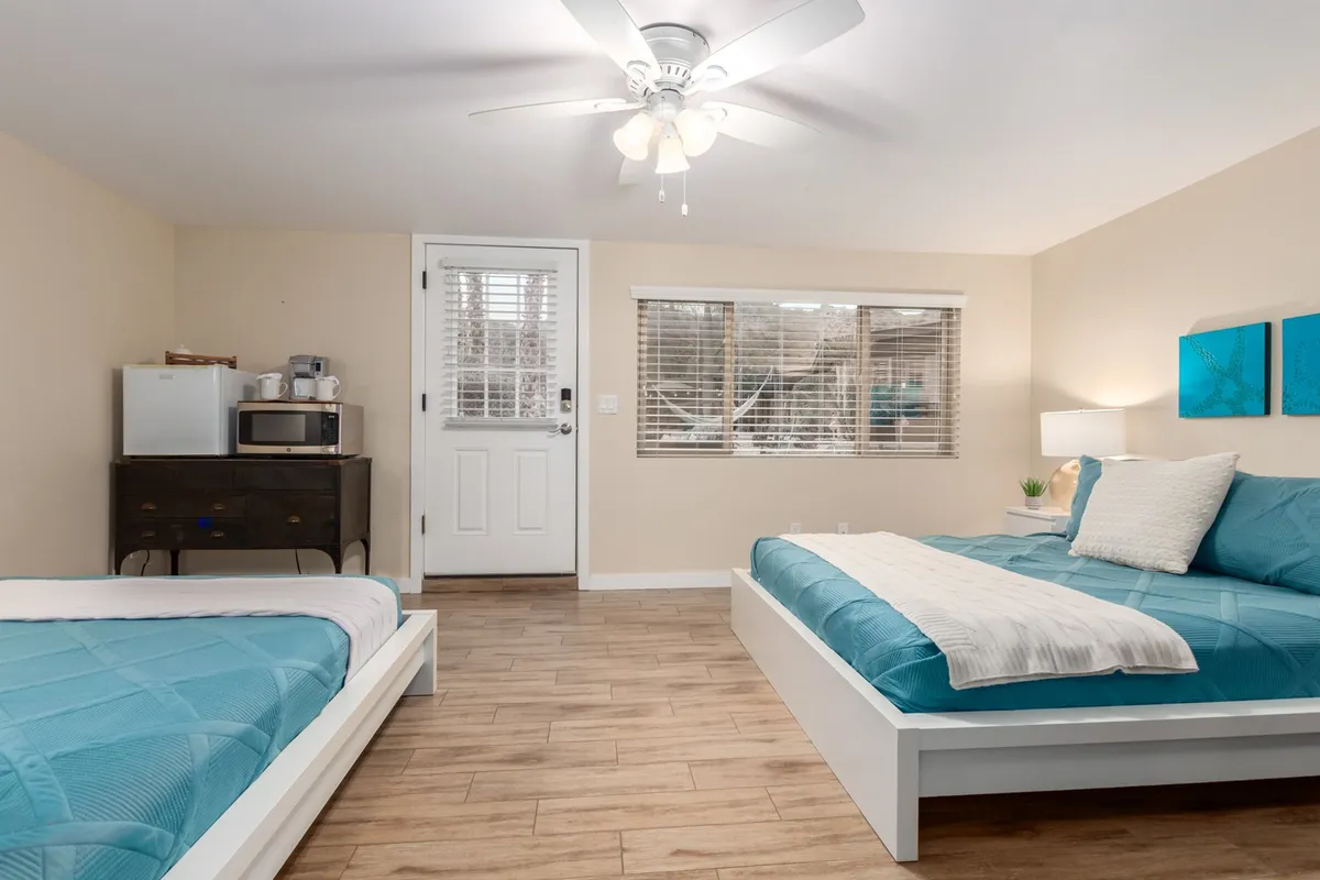 A comfortable casita bedroom with two beds featuring turquoise bedding, decorative blue wall art, ceiling fan, and a kitchenette area with a microwave and fridge. The setup is perfect for light meals or snacks and offers privacy and convenience for guests seeking a separate retreat.