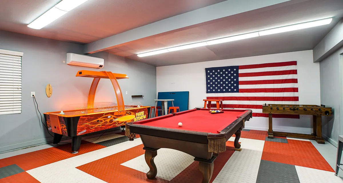 A spacious interior game room with a pool table, air hockey table, and foosball table arranged for group entertainment. An American flag decorates the wall, contributing to a playful, welcoming vibe. Friends can be seen engaging in games and conversation.