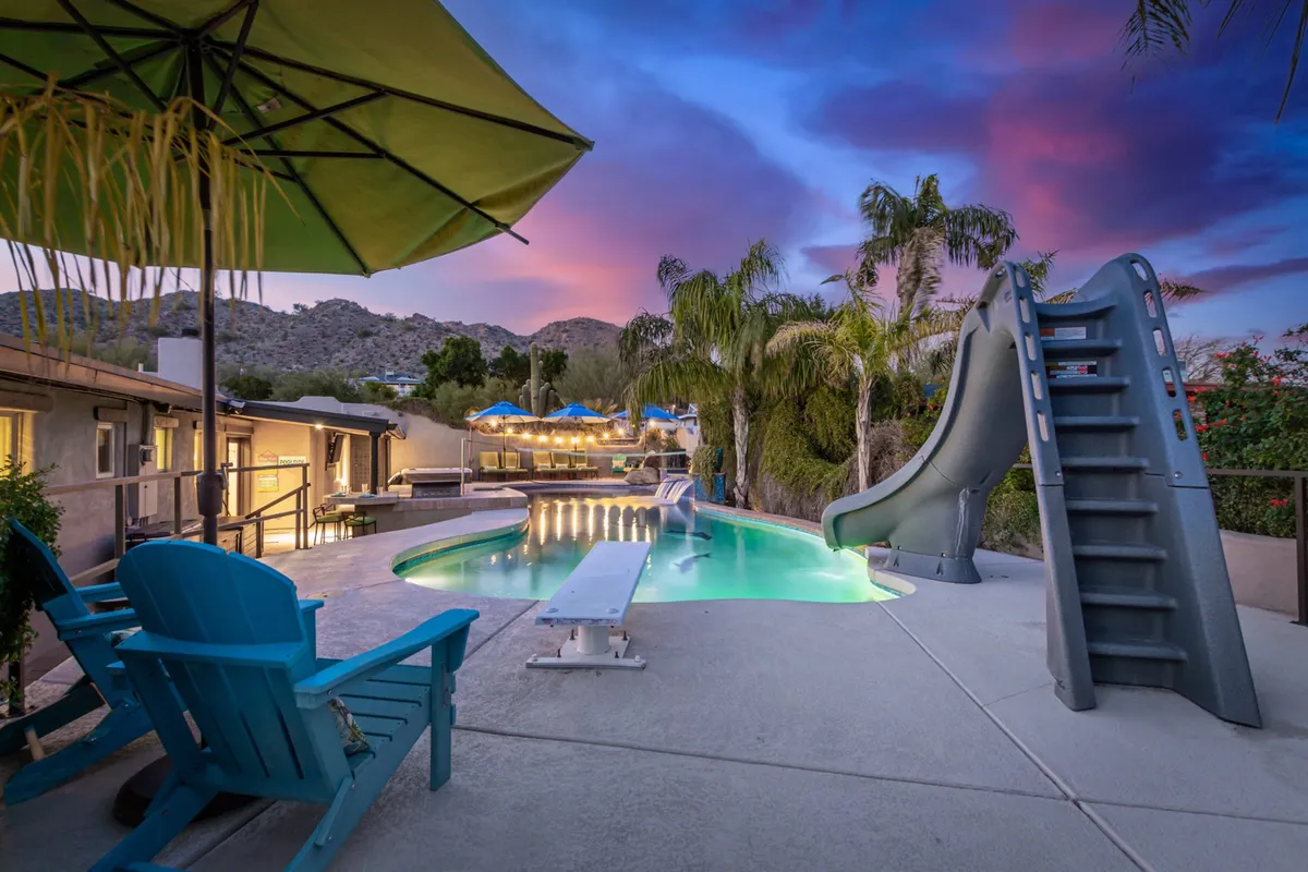 This image captures a sparkling swimming pool with an inviting water slide, surrounded by comfortable outdoor seating and umbrellas, set within lush landscaping. The scene is lively yet relaxing, suggesting both energetic pool play and sun-soaked lounging for all ages.