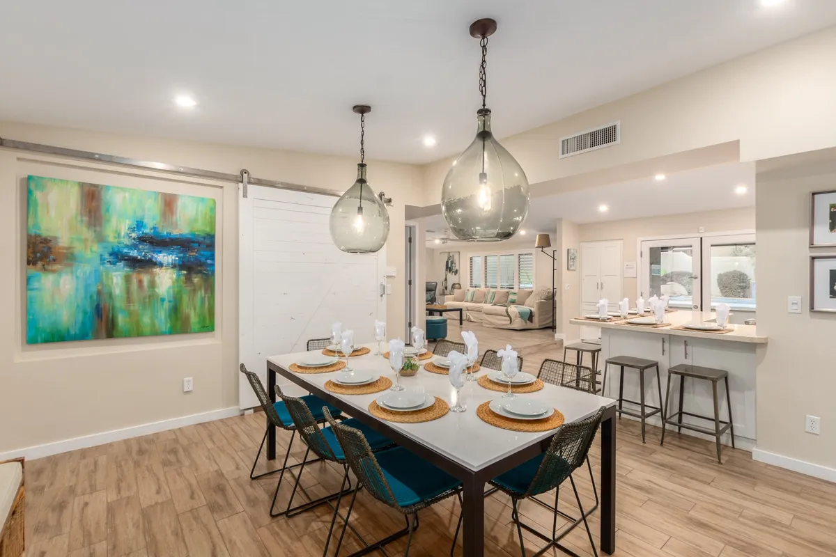 A stylish dining area with a large table set for eight, contemporary light fixtures overhead, a sliding barn door, and a striking abstract painting on the wall. The space is bright, modern, and purposely arranged for group dining or games.