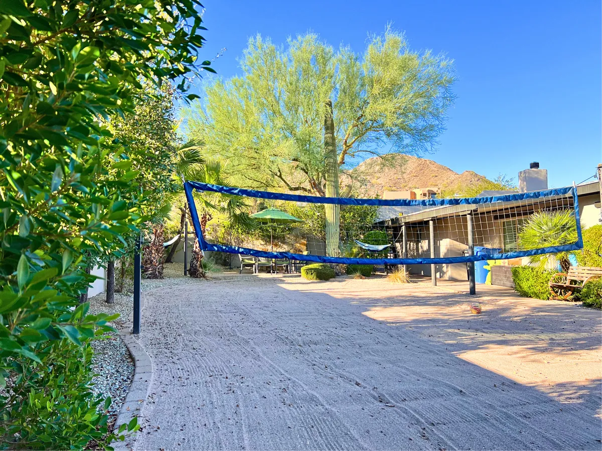 This exterior image captures a lively backyard scene with a volleyball net set up on green grass, surrounded by lush foliage, a mountain view in the distance, and comfortable outdoor seating. The setup hints at the possibility of casual games and relaxing nature-filled moments.