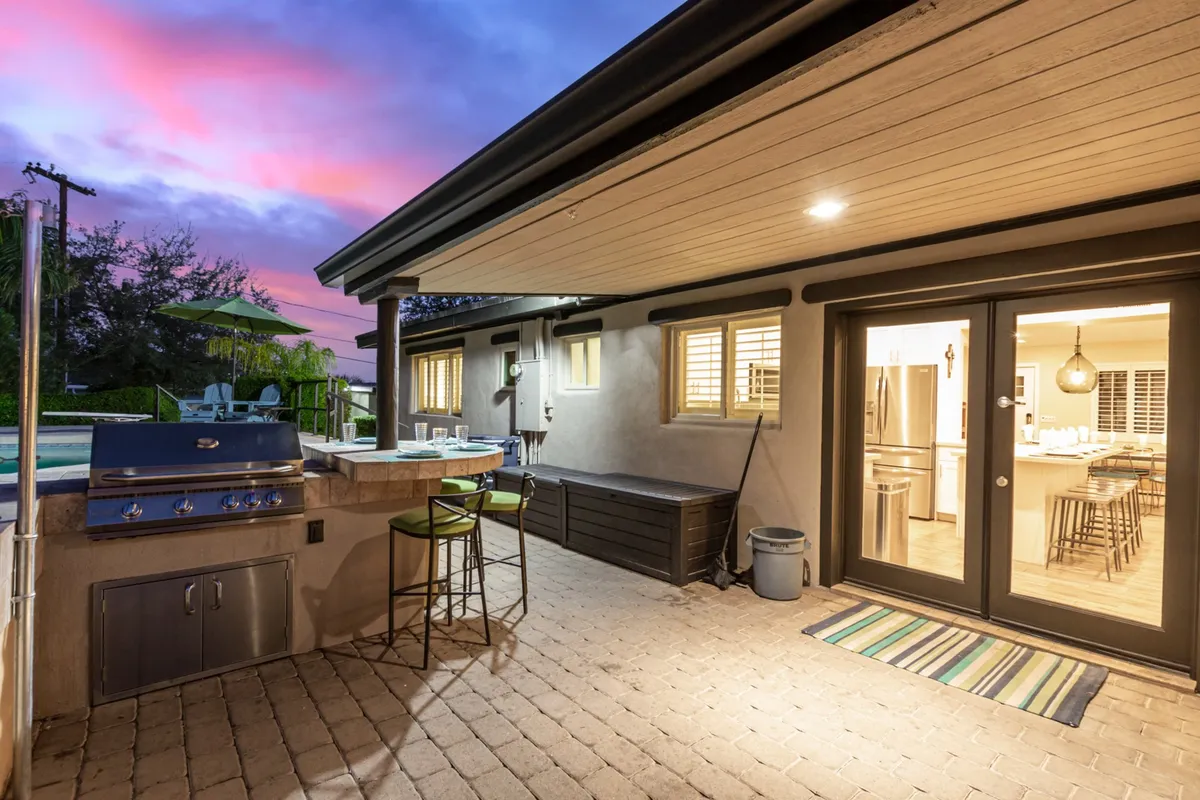 This exterior shot showcases an outdoor grill station with bar stools and a dining area, set against a sunset sky. The setup is ideal for barbecues, outdoor meals, and socializing with friends and family, echoing the convenience of nearby grocery stores and the option for home-cooked meals.