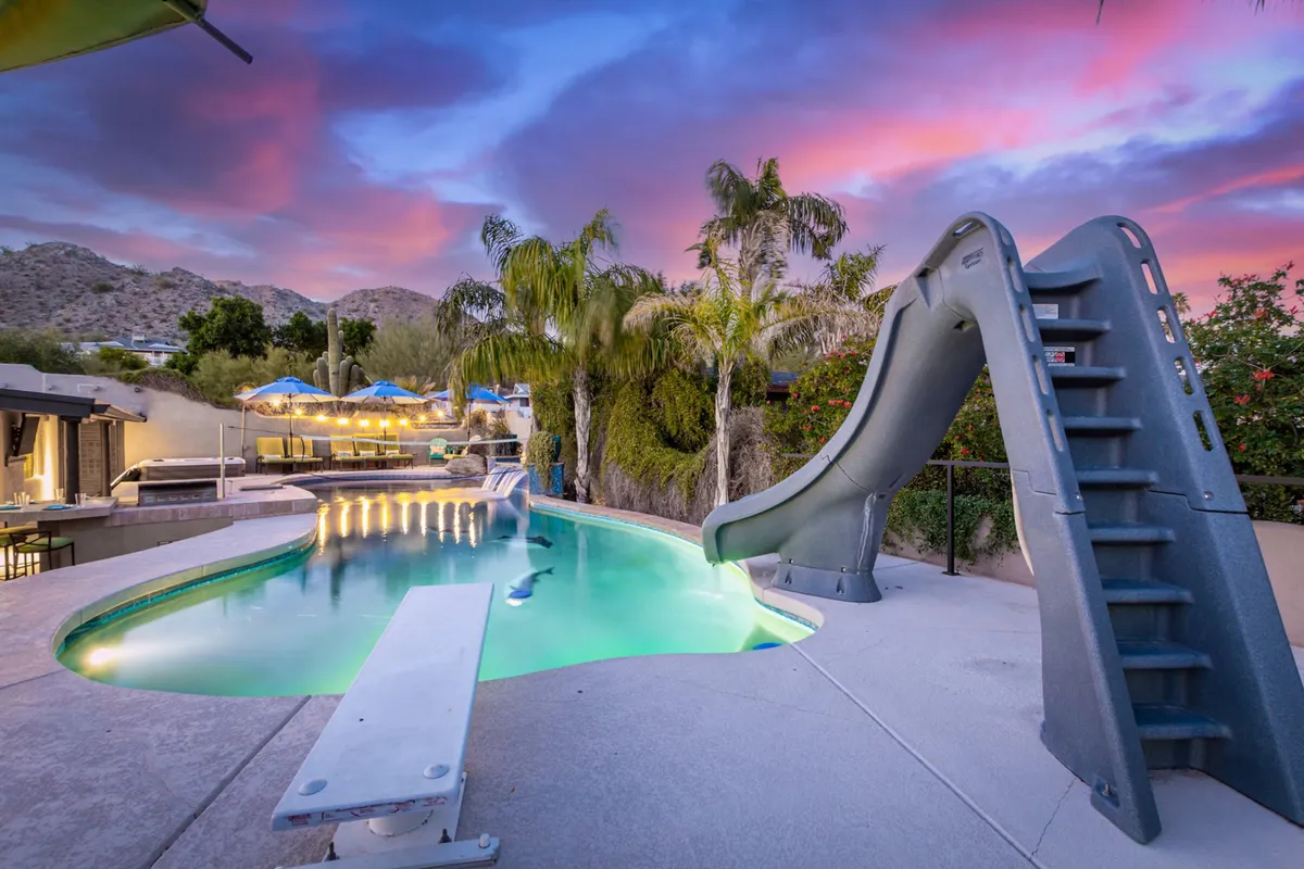 This image features a vibrant swimming pool area with a water slide, surrounded by palm trees and an adjacent outdoor dining space. The scene is lively and inviting, ideal for both relaxation and fun-filled family gatherings. The setting is bathed in warm light, suggesting an all-season appeal, whether for swimming in summer or enjoying the view year-round.