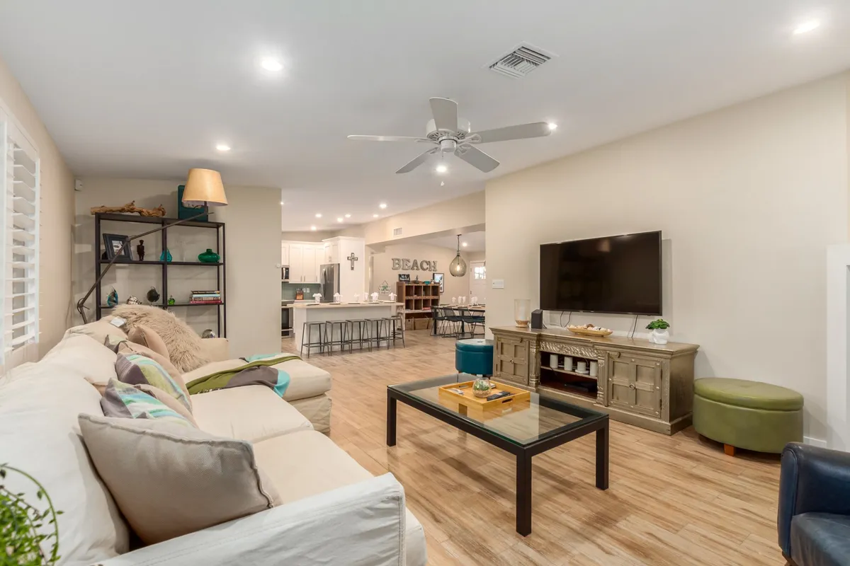 A cozy, open living room with a plush sofa, coffee table, and large television, seamlessly connected to an open kitchen area in the background. The space is filled with natural light from oversized windows, creating a relaxed and sociable environment.