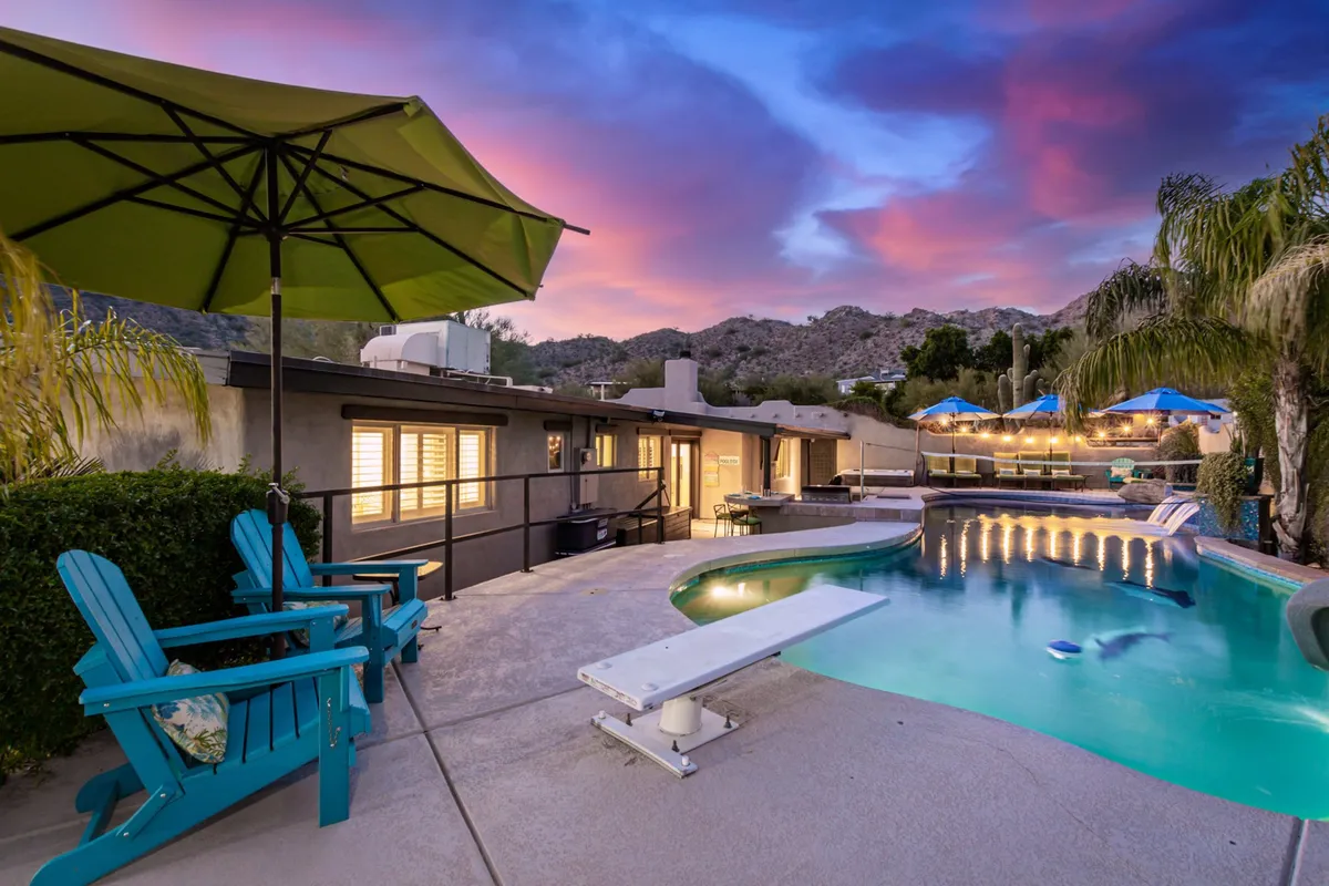 A serene swimming pool scene at twilight, framed by adirondack chairs, an umbrella, mountain views, and twinkling lights overhead. The space is designed for relaxation and group enjoyment, with the tranquil pool reflecting the dramatic Arizona sunset and inviting guests to swim, dine, or unwind together.