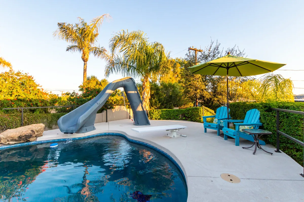 A vibrant backyard scene featuring a sparkling pool with a water slide, surrounded by lounge chairs, umbrellas, and palm trees. The image captures both the pool area and the lounging deck, with guests swimming, sunbathing, and relaxing under the shade of umbrellas, all beneath a colorful sunset sky.