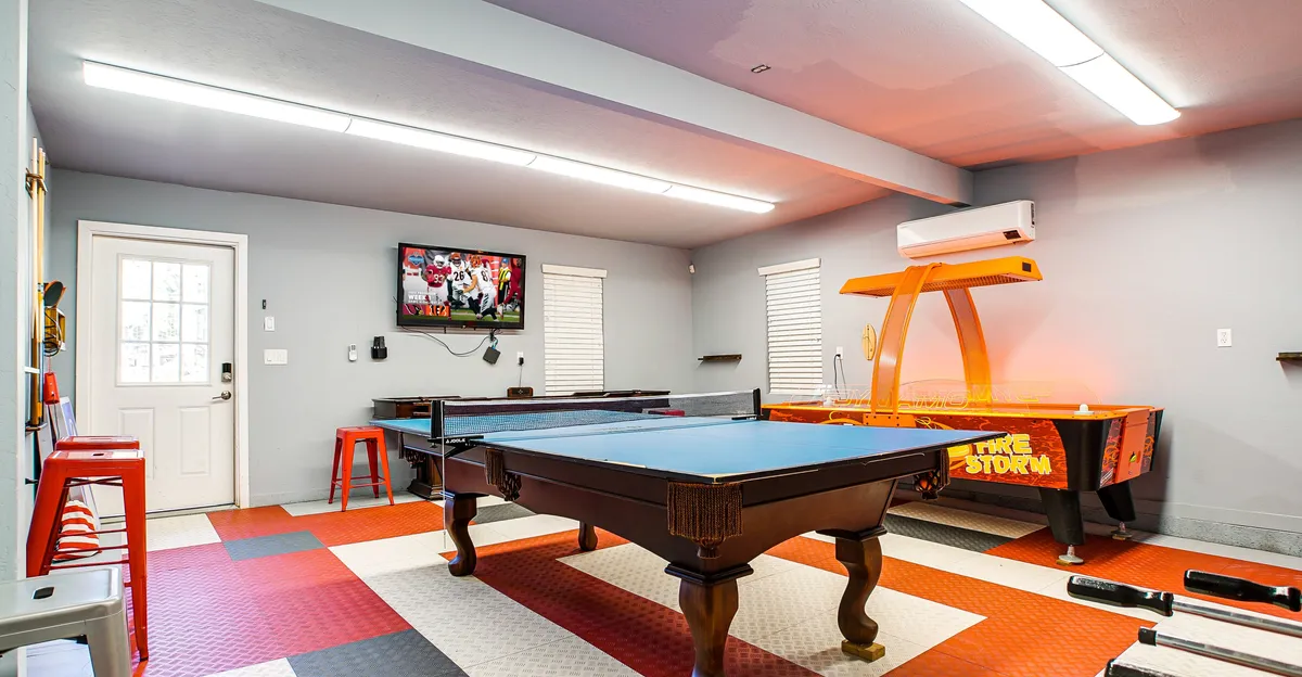 A lively interior game room featuring a pool table at the center, an arcade game in one corner, and a large television mounted on the wall. The room is set up for both playing and watching sports, with ample open space for socializing. The lighting and decor contribute to a fun, energetic atmosphere, ideal for group activities and friendly competitions.