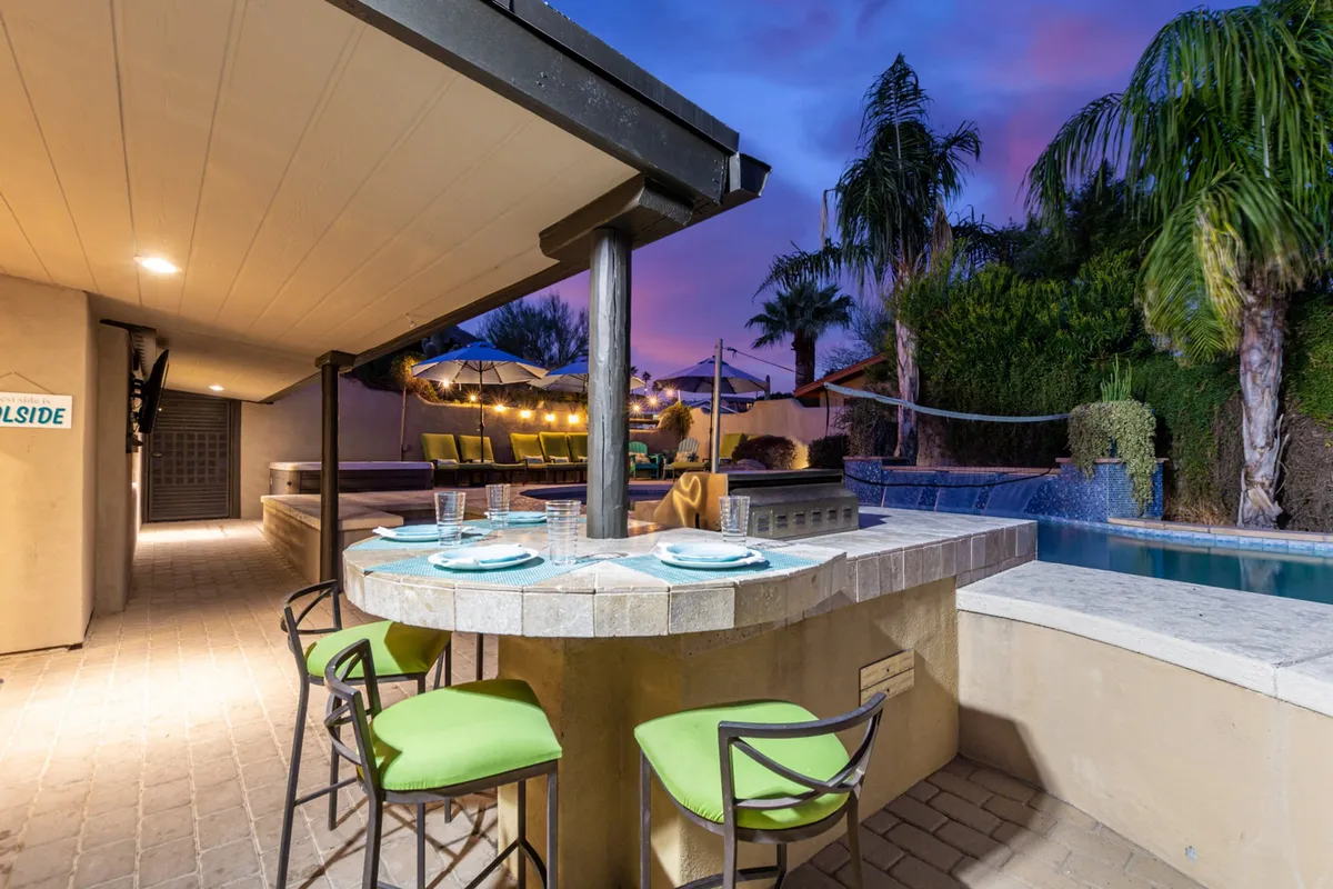A stylish outdoor bar area sits adjacent to a sparkling pool, framed by swaying palm trees and decorative string lighting. The bar is equipped with comfortable stools and a countertop perfect for evening cocktails, while the pool area offers additional lounging opportunities for groups. The setting glows with warm, ambient lighting as dusk approaches, inviting guests to gather and socialize.