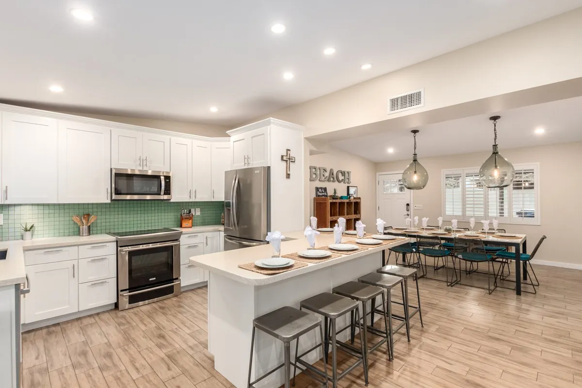 This image displays an expansive open-plan living and dining area, featuring a prominent kitchen island and a large dining table. The space is styled with inviting beach-themed decor, ample seating, and bright natural light, reflecting a welcoming and communal atmosphere.
