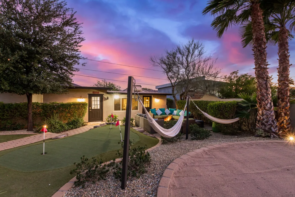 A lush backyard scene featuring hammocks strung between palm trees, a putting green for friendly golf competitions, and cozy fire pit seating. The space is designed for relaxation, play, and evening gatherings under the stars.