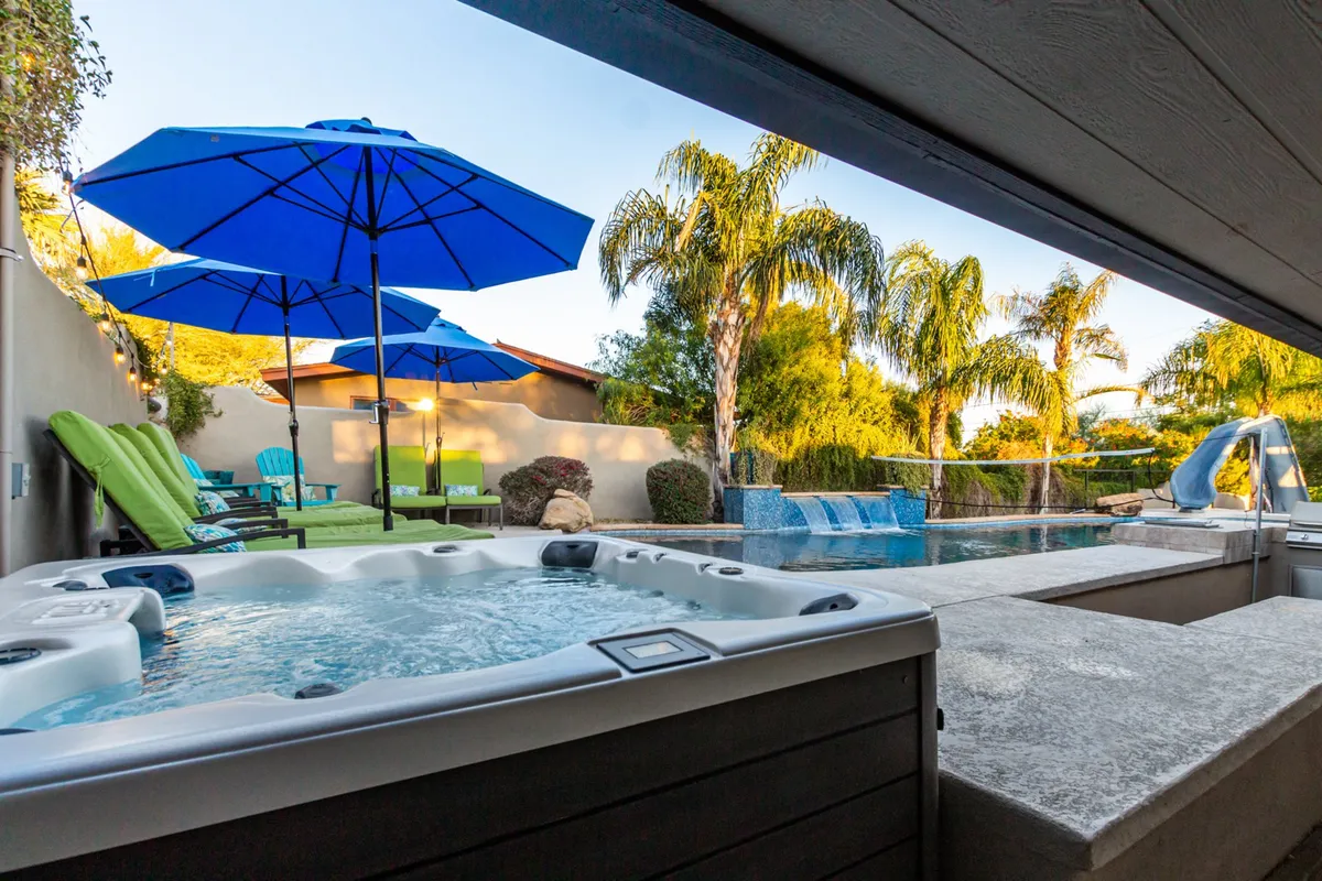 This image focuses on the hot tub adjacent to the pool, with blue umbrellas, plush lounge chairs, and tropical palm trees creating a tranquil oasis. The scene exudes relaxation and luxury, with guests enjoying the hot tub and sunbathing nearby.
