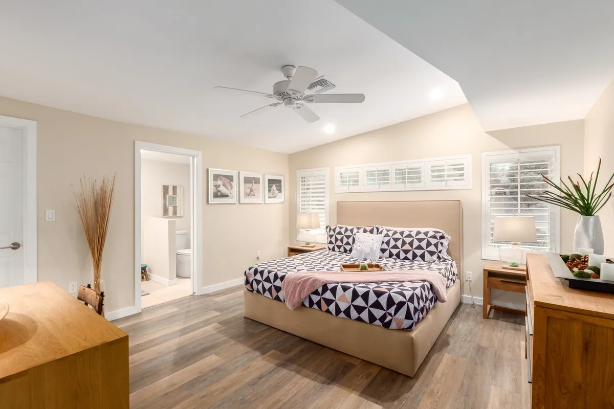 A tranquil bedroom with a large, comfortable bed dressed in patterned bedding, wooden dresser, framed art on the wall, and a potted plant for a homely touch. The room is bathed in natural light, creating a serene and cozy ambiance ideal for relaxation and privacy.