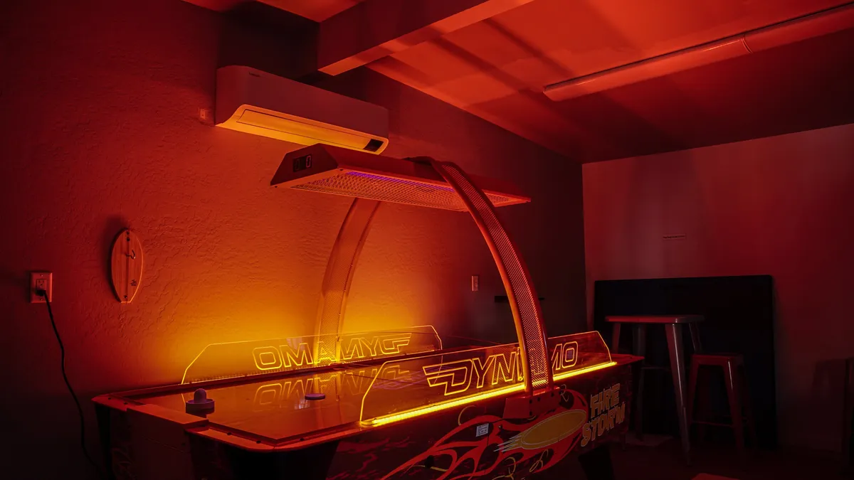Inside the fully equipped game room, a neon-lit air hockey table sits beneath ambient lighting, with a wall-mounted air conditioner visible. The mood is energetic and fun, perfect for cooling off and spending quality time indoors during the summer or evenings.