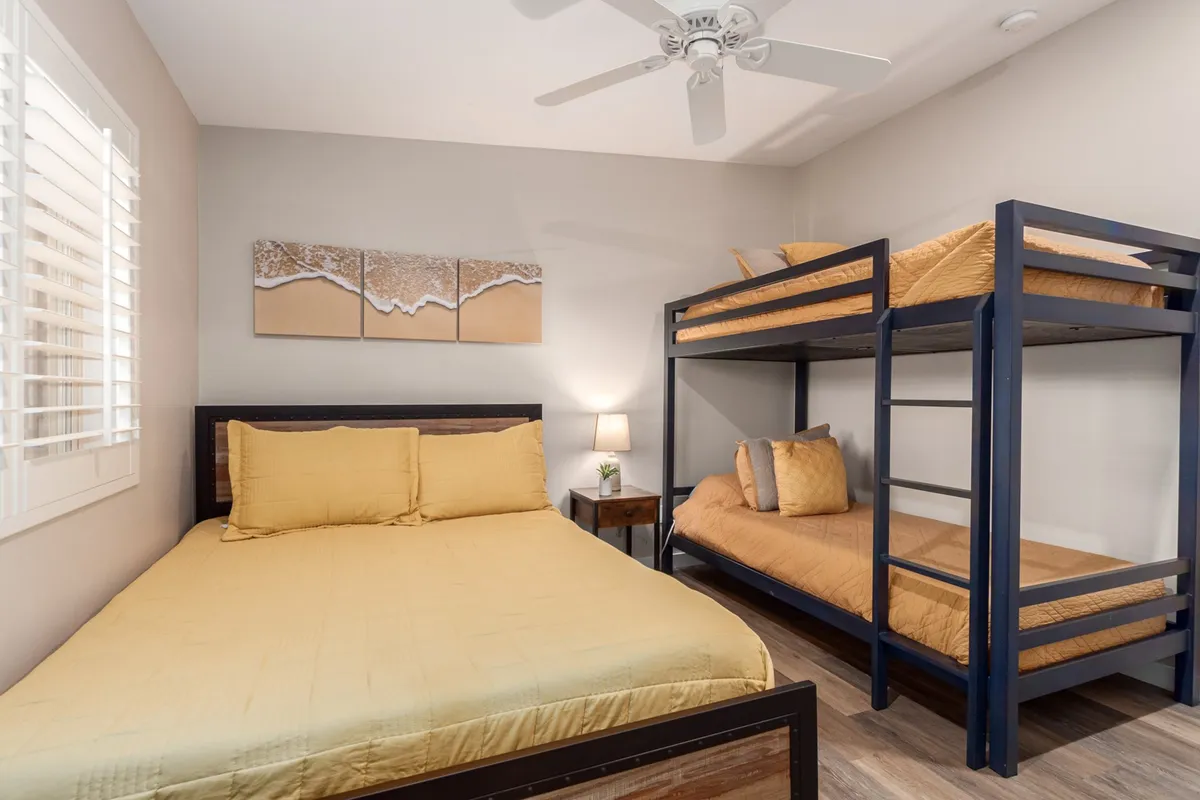 This inviting bedroom scene features a bunk bed and a queen bed, adorned with soft bedding and gentle lighting from a table lamp. Artwork on the wall adds a personal touch, while the window allows natural light to fill the space. The room is spacious, cozy, and perfectly suited for families or groups with children.
